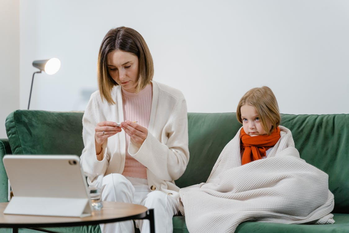 An image of a mother sitting on a couch with her sick daughter while checking her temperature during an online medical appointment